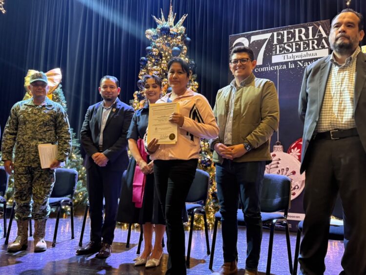 Un grupo de seis personas posa en un escenario decorado con árboles y adornos navideños, mientras una mujer sostiene un certificado.