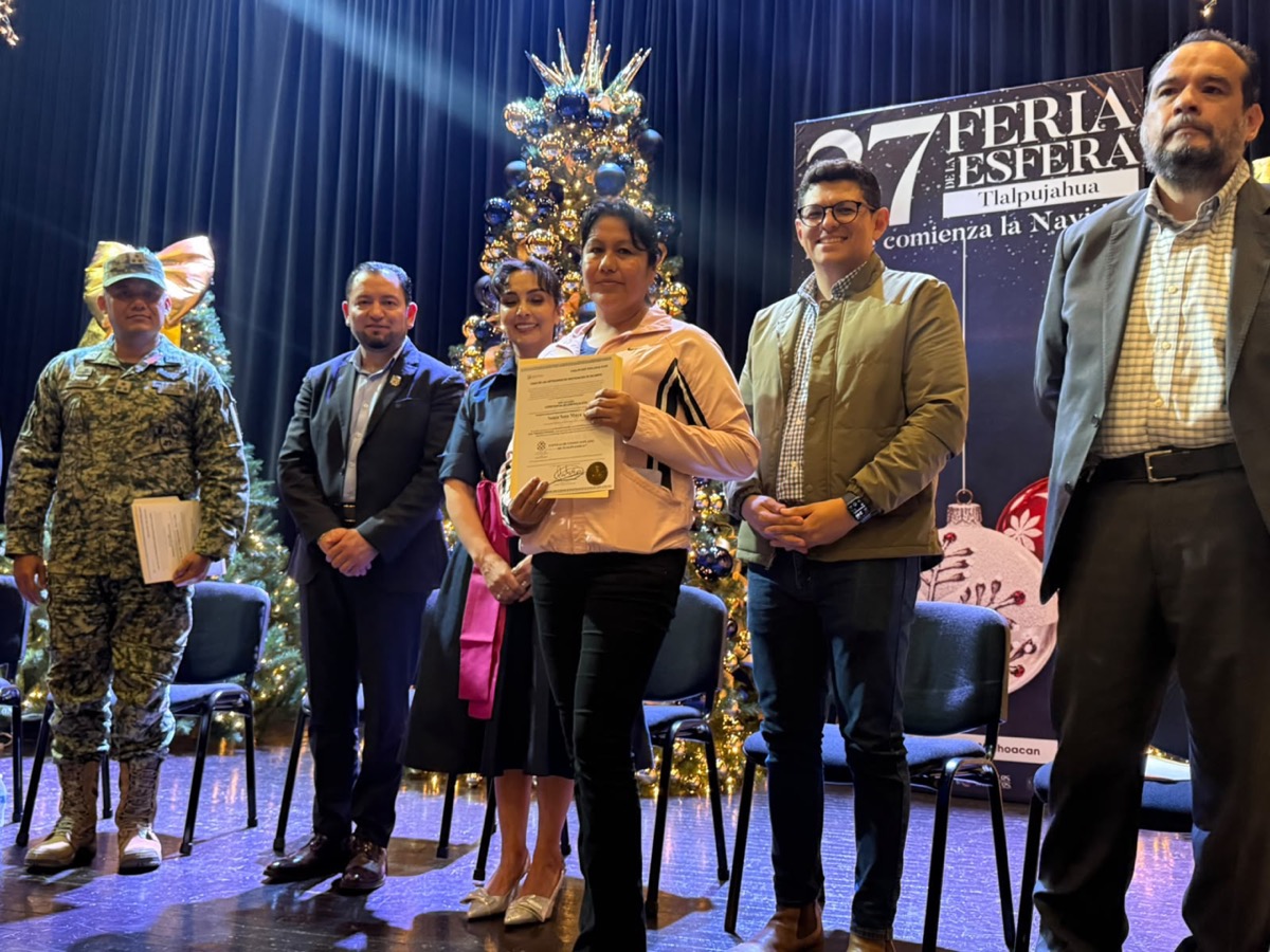 Un grupo de seis personas posa en un escenario decorado con árboles y adornos navideños, mientras una mujer sostiene un certificado.