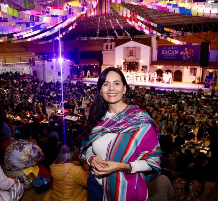 Una mujer sonriente con un rebozo colorido está en un auditorio lleno de gente, frente a un escenario decorado con banderas de colores.