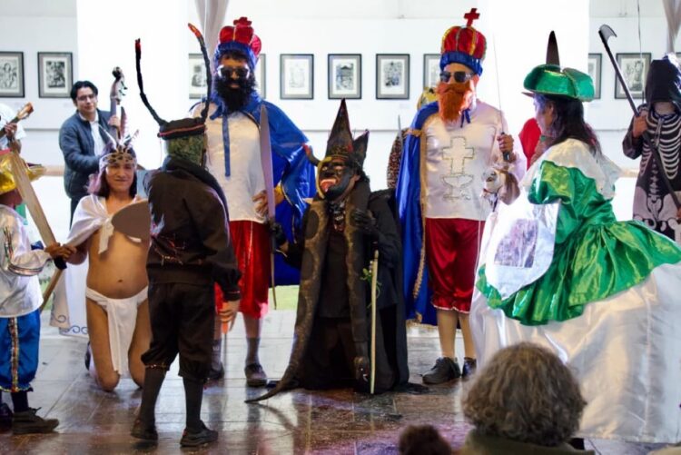 Un grupo de personas disfrazadas con trajes coloridos y máscaras participa en una representación teatral en un espacio interior.
