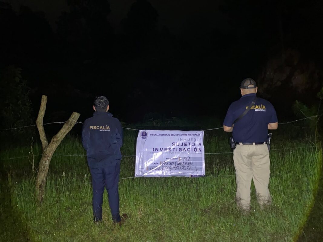 Dos agentes de la Fiscalía de Michoacán observan un cartel de investigación colocado en una cerca durante la noche.