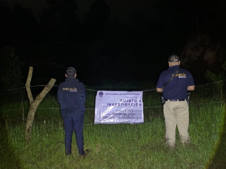 Dos agentes de la Fiscalía de Michoacán observan un cartel de investigación colocado en una cerca durante la noche.
