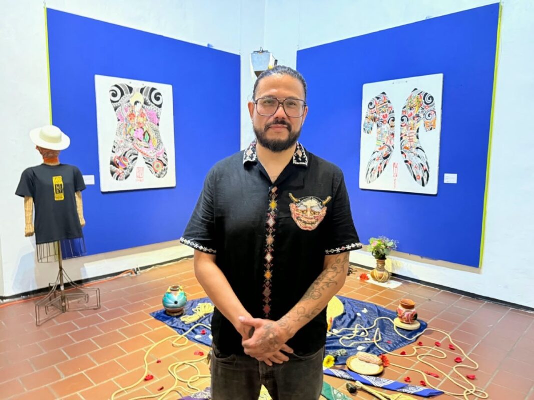 Un hombre con barba y gafas está de pie en una galería de arte con murales coloridos y objetos en el suelo.
