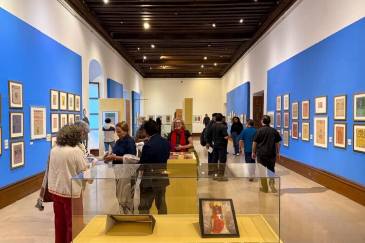 Personas observan cuadros y exhibiciones en una galería de arte con paredes azules.