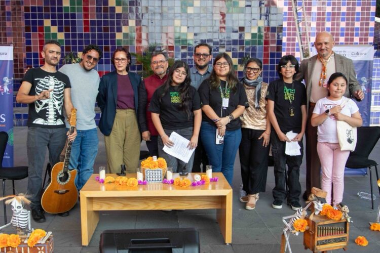 Un grupo de personas posa sonriente frente a un muro de mosaicos coloridos, con una mesa decorada con flores de cempasúchil y figuras de esqueletos al frente.