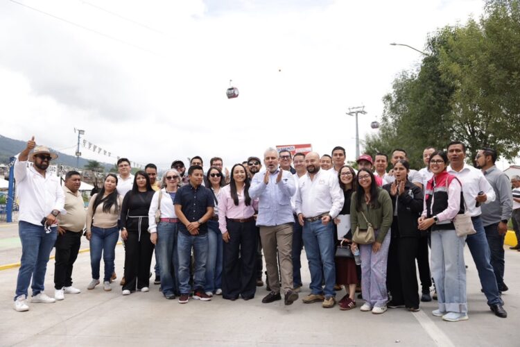 Un grupo de personas posa sonriente al aire libre sobre una calle, con árboles y teleféricos al fondo.