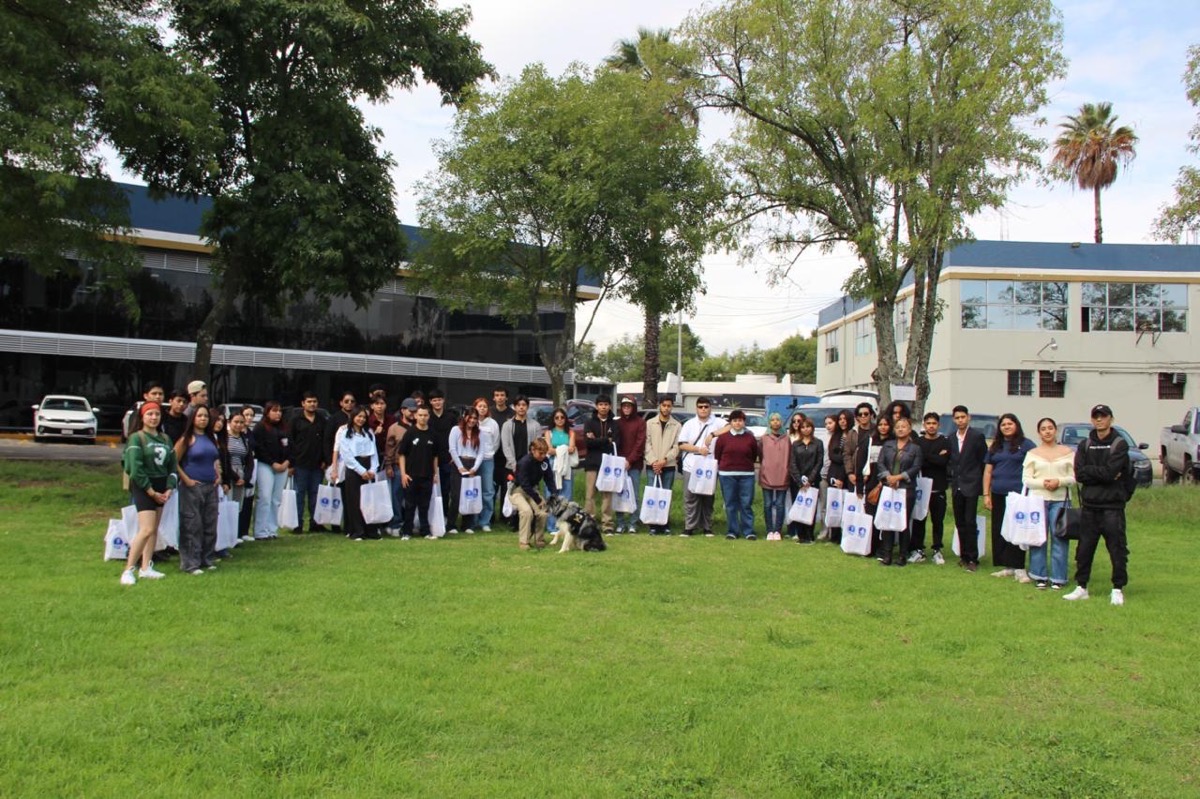 Un grupo de personas está reunido al aire libre en un césped con árboles, sosteniendo bolsas blancas, mientras un perro se encuentra al frente.