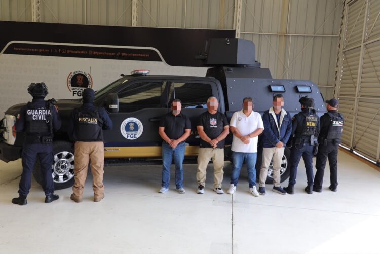 Varios agentes armados escoltan a cuatro hombres esposados frente a un vehículo blindado dentro de un hangar.