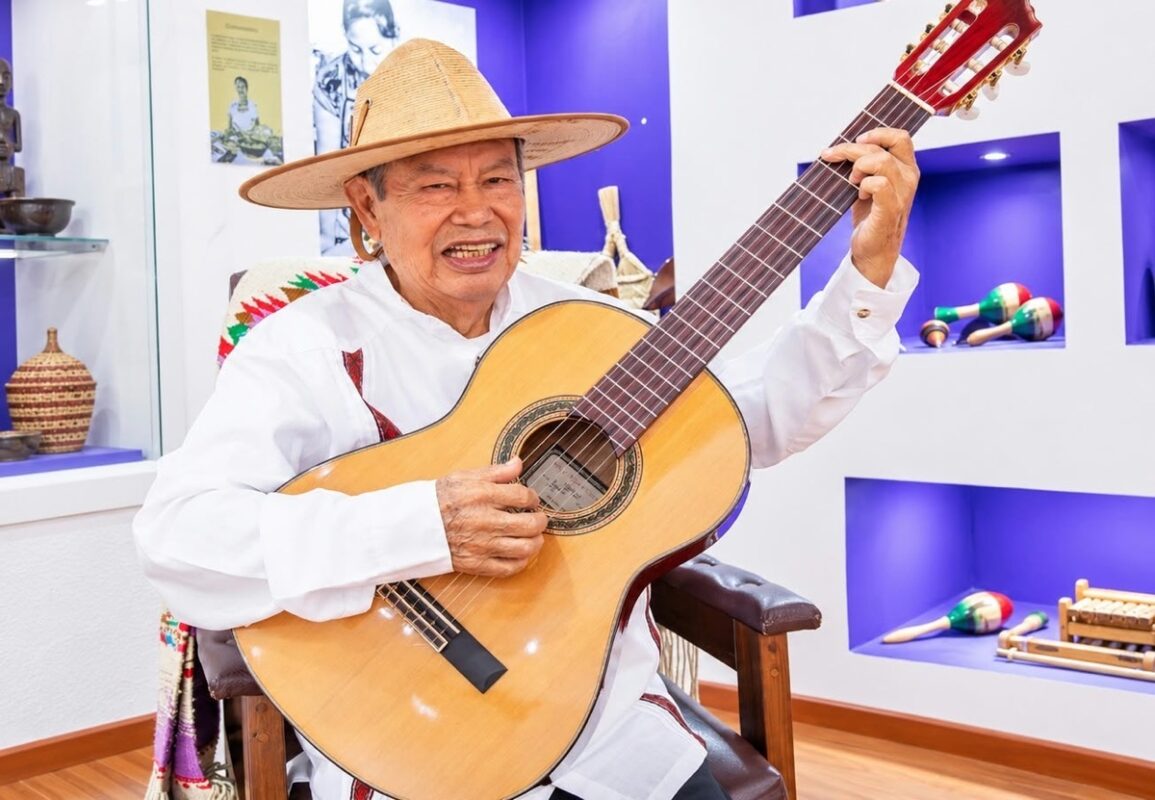 Un hombre mayor con sombrero toca una guitarra mientras está sentado en una habitación decorada con instrumentos musicales.