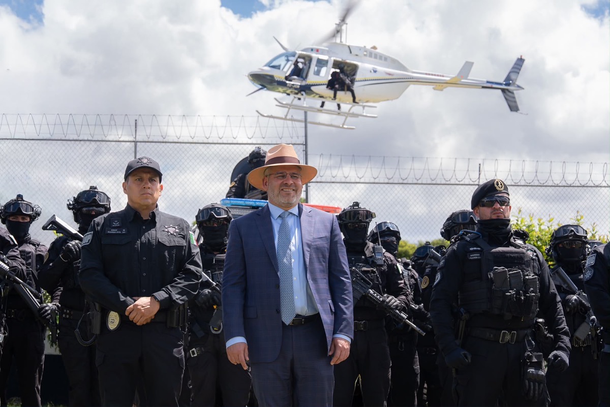 Un grupo de policías fuertemente armados posa junto a un hombre con traje y sombrero frente a una cerca, mientras un helicóptero vuela detrás.