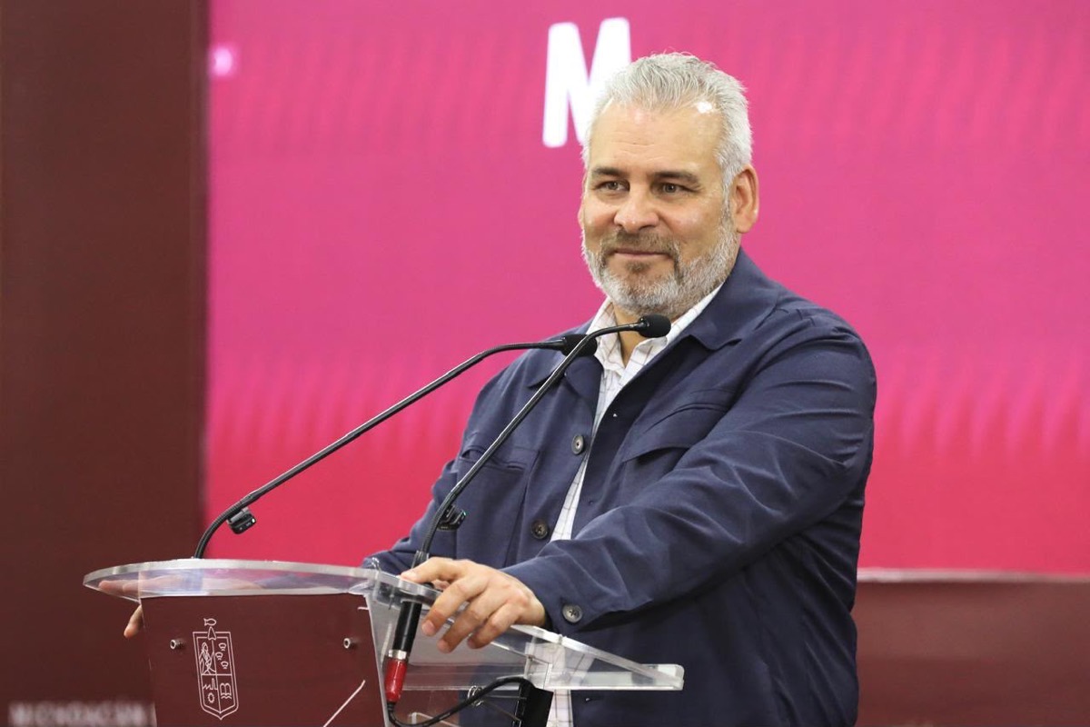 Un hombre de cabello canoso y barba está hablando en un podio con micrófonos frente a un fondo rosa.