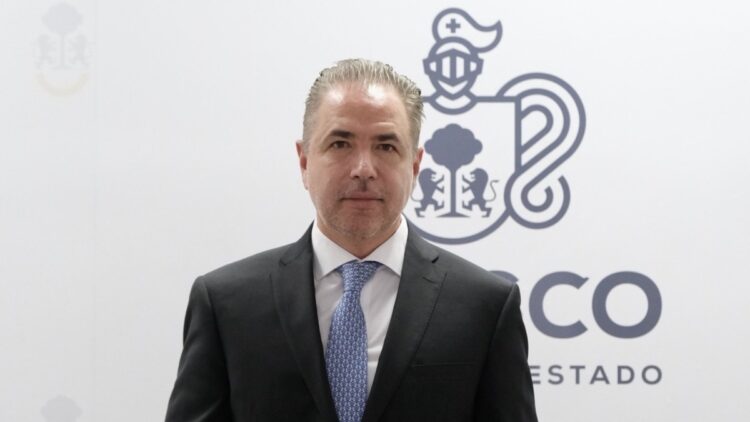Un hombre con traje y corbata posa frente a un fondo blanco con un logotipo institucional.