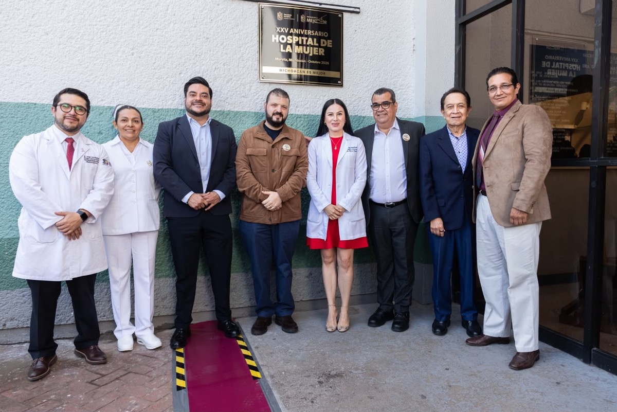 Un grupo de ocho personas, algunas con bata médica y otras con ropa formal, posa frente a una placa conmemorativa en la entrada de un edificio.