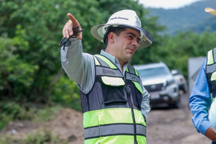 Un hombre con casco y chaleco reflectante señala hacia un lado mientras está al aire libre junto a otra persona parcialmente visible.