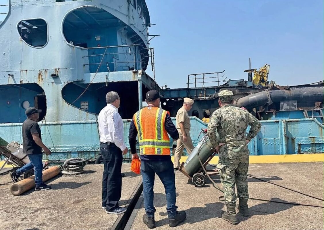 Varias personas, incluyendo personal militar y civil, observan un barco azul desde un muelle bajo un cielo despejado.
