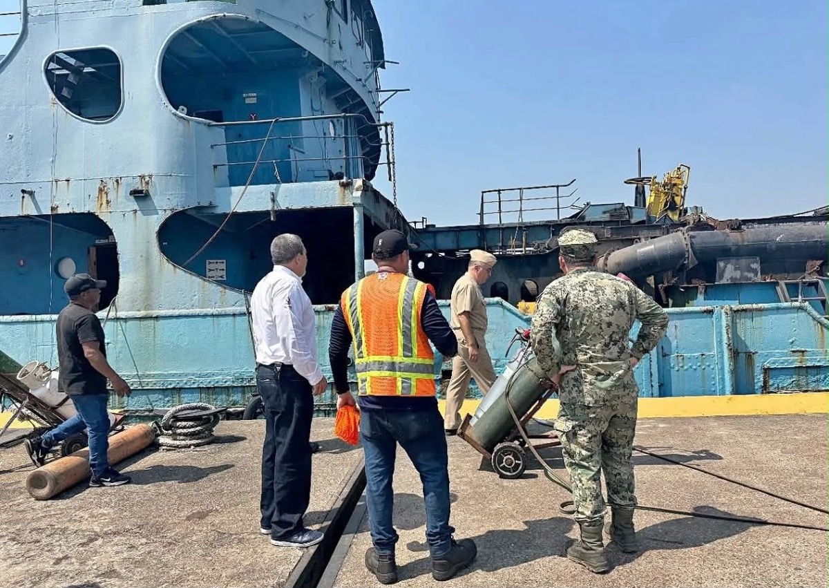 Trabajadores y militares inspeccionan una barcaza azul dañada, con equipo de protección y herramientas en el muelle.