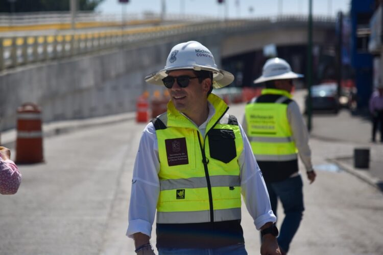 Un hombre con casco y chaleco reflectante sonríe mientras supervisa una obra vial al aire libre.