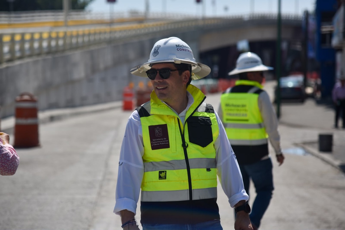 Un hombre con casco y chaleco reflectante sonríe mientras supervisa una obra vial al aire libre.