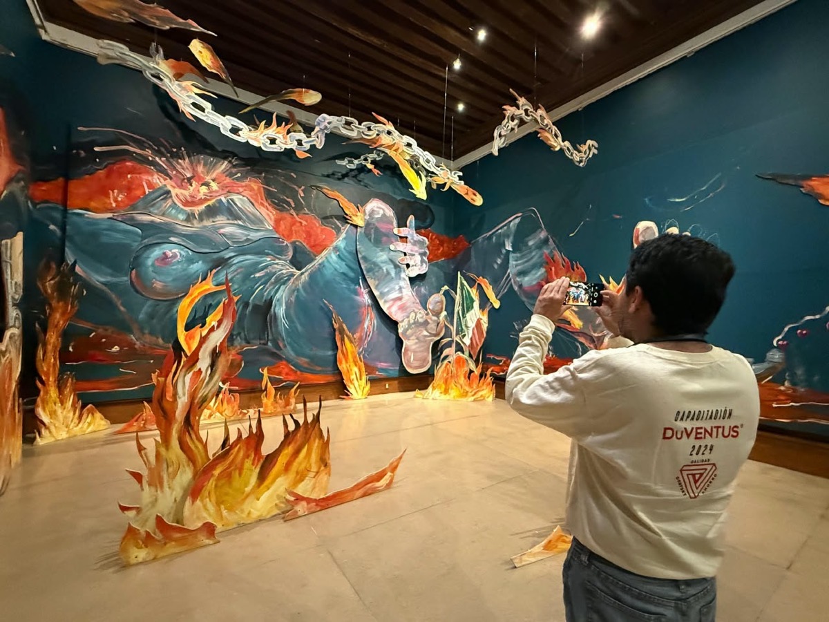 Un hombre toma una foto de un mural colorido que representa figuras en llamas dentro de una galería.