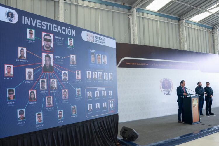 Un funcionario presenta un organigrama de investigación con fotos de personas detenidas en una conferencia de prensa dentro de un recinto oficial.