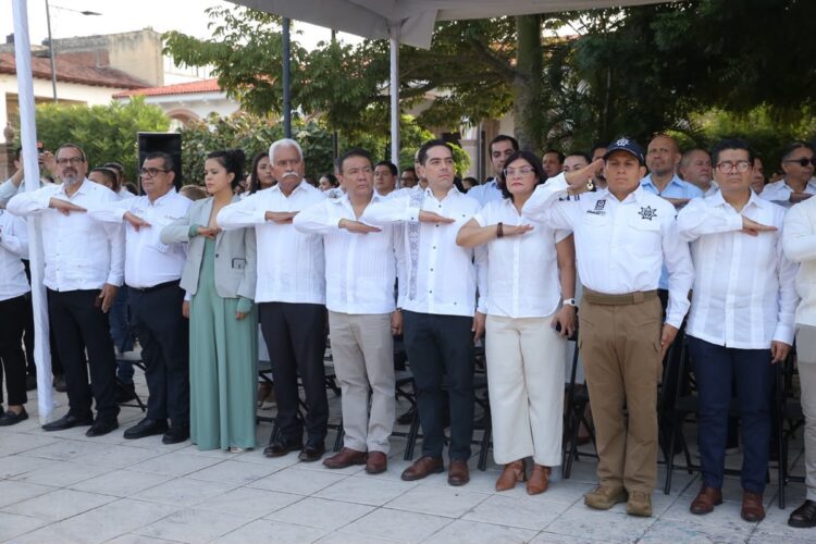 Un grupo de personas vestidas de blanco y beige hace un saludo cívico al aire libre bajo una carpa.