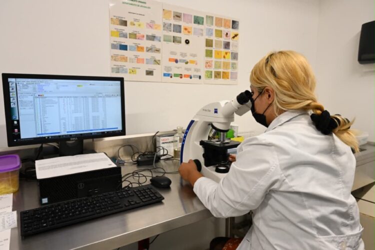 Una persona con bata de laboratorio y mascarilla observa un microscopio junto a una computadora en un laboratorio.