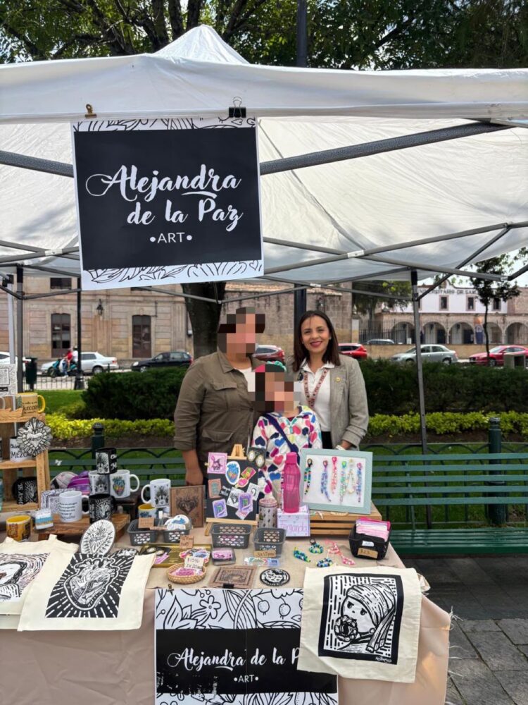 Tres personas están detrás de un puesto al aire libre que exhibe artesanías y productos decorativos bajo un toldo blanco.