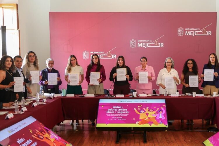 Un grupo de personas está de pie en una sala sosteniendo documentos frente a una mesa con un fondo rojo que dice “Michoacán es Mejor”.