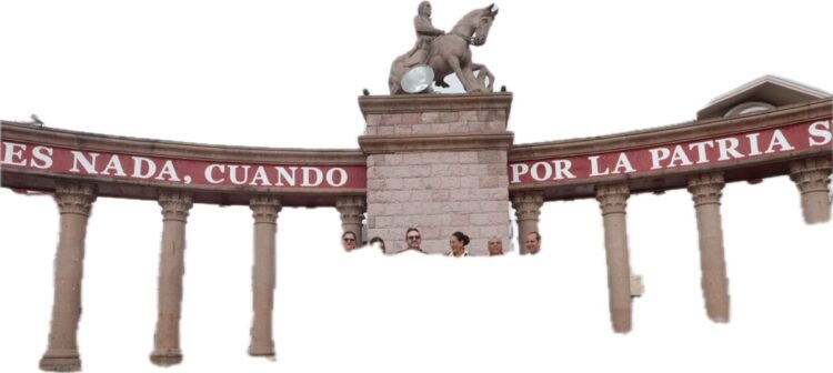 Un monumento con columnas y una estatua ecuestre, con personas asomándose entre las columnas y un letrero rojo con texto blanco parcialmente visible.
