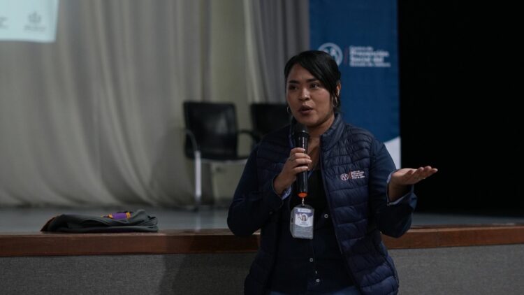 Una mujer sostiene un micrófono y habla en un auditorio frente a un escenario con sillas vacías.