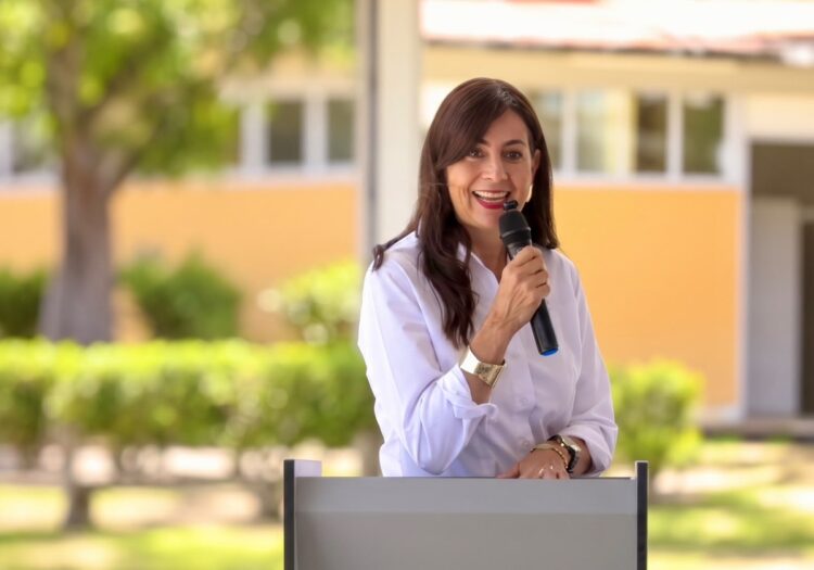 Una mujer sonriente con camisa blanca habla por un micrófono detrás de un podio al aire libre.