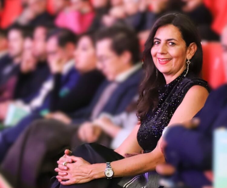 Una mujer sonriente vestida de negro está sentada entre otras personas en un auditorio.