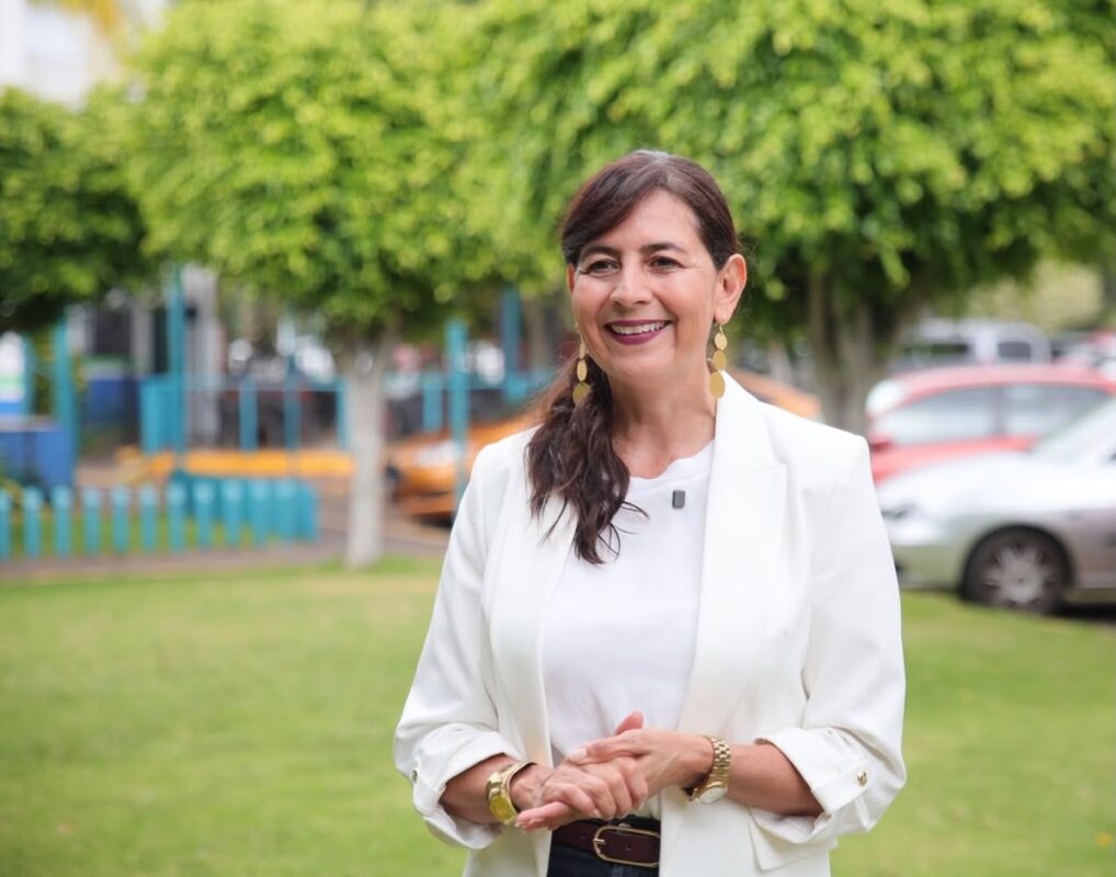 Una mujer sonriente con chaqueta blanca está de pie en un parque con árboles y autos al fondo.