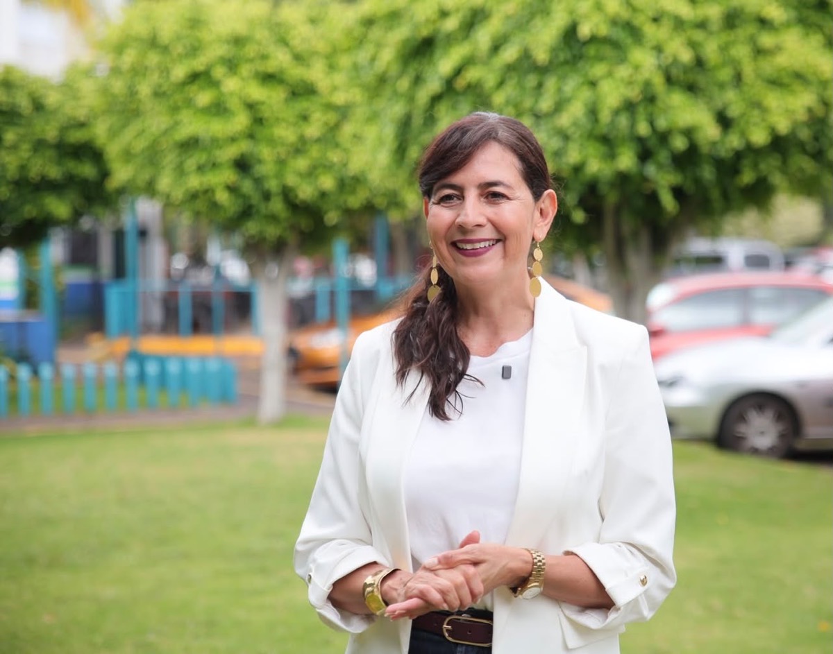 Una mujer sonriente con chaqueta blanca está de pie en un parque con árboles y autos al fondo.
