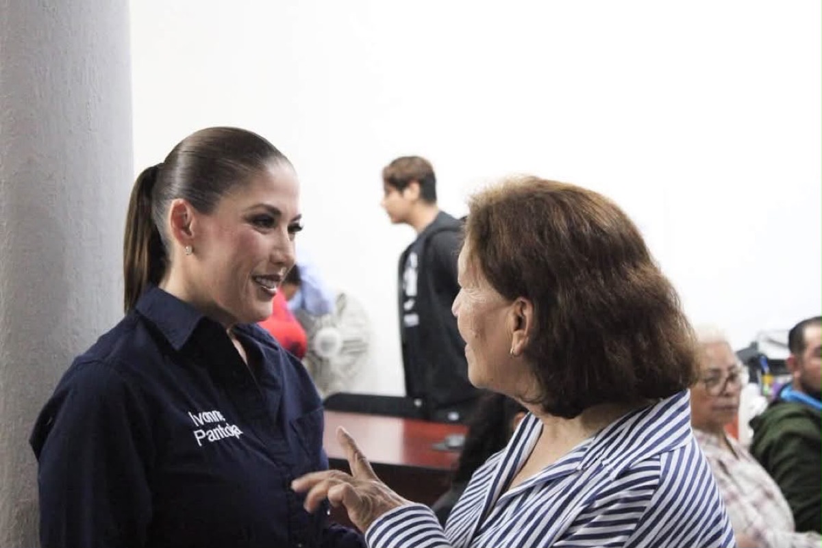 Dos mujeres conversan en un interior, mientras otras personas están sentadas al fondo.