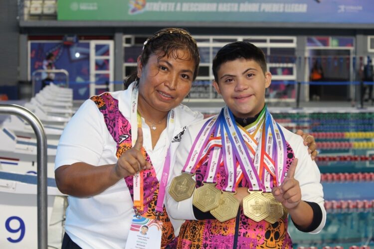 Dos personas sonríen junto a una piscina, y una de ellas lleva varias medallas colgadas en el cuello.