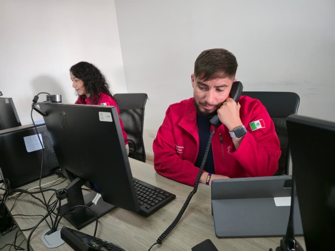 Dos personas con chaquetas rojas trabajan en escritorios con computadoras, una usando un teléfono y la otra escribiendo.