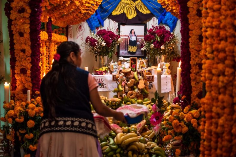 Mujer coloca ofrendas de frutas y pan en un altar purépecha con flores de cempasúchil y velas encendidas.