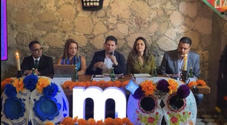Cinco personas están sentadas detrás de una mesa decorada con calaveras y flores, mientras una de ellas habla con un micrófono.