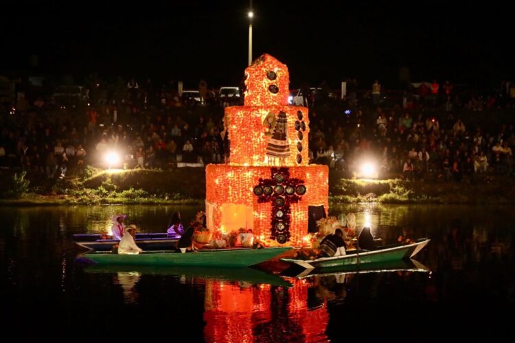 Un altar iluminado flota sobre el agua de noche, rodeado de personas en pequeñas lanchas y con un público observando desde la orilla.