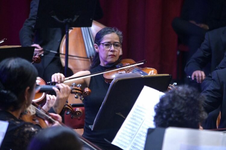 Una mujer toca el violín en una orquesta rodeada de otros músicos y partituras.