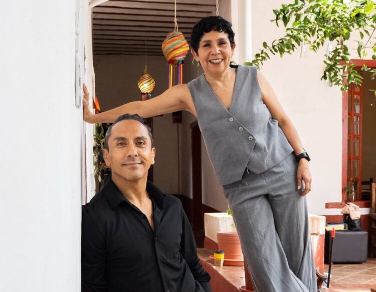 Un hombre con camisa negra y una mujer con traje gris sonríen en un patio decorado con macetas y lámparas colgantes de colores.