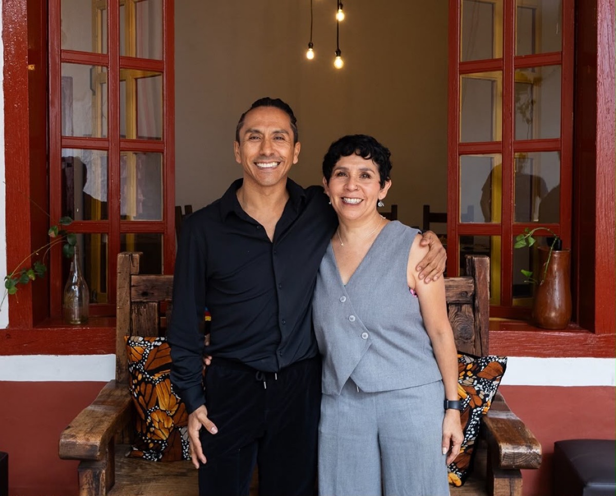 Dos personas sonrientes posan abrazadas frente a una ventana con marcos rojos, sentadas en un banco de madera con cojines de mariposas.