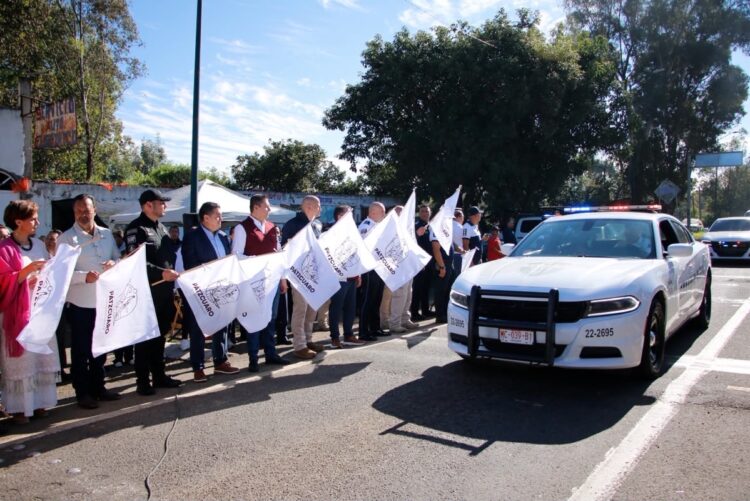Un grupo de personas sostiene banderas blancas mientras un auto de policía blanco pasa frente a ellos en una calle soleada.