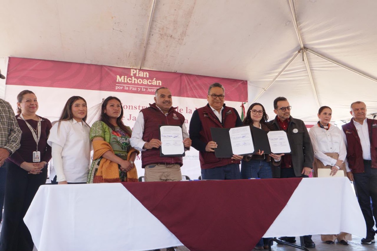 Un grupo de personas posa bajo una carpa mientras algunas sostienen documentos firmados frente a una mesa con mantel blanco y burdeos.