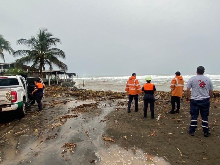Varias personas con chalecos y uniformes observan una playa cubierta de escombros junto al mar agitado.