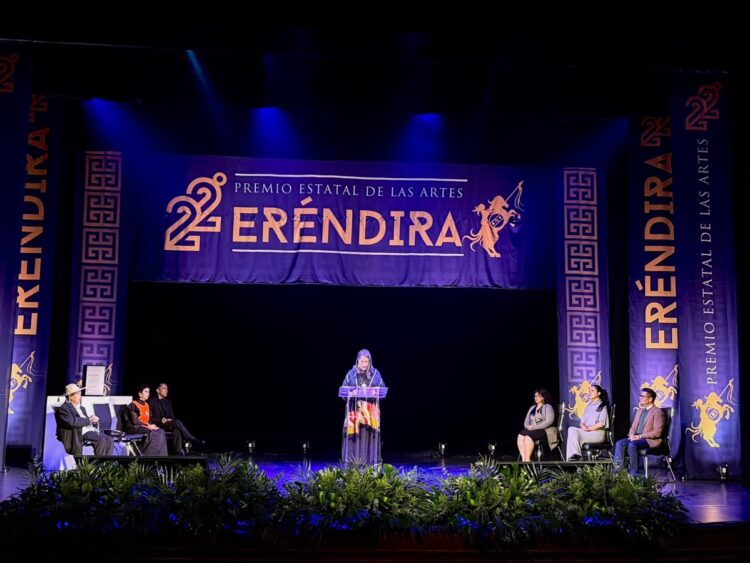 Un escenario con una persona hablando en un podio, flanqueada por varias personas sentadas y decoraciones que dicen “Premio Estatal de las Artes Eréndira”.