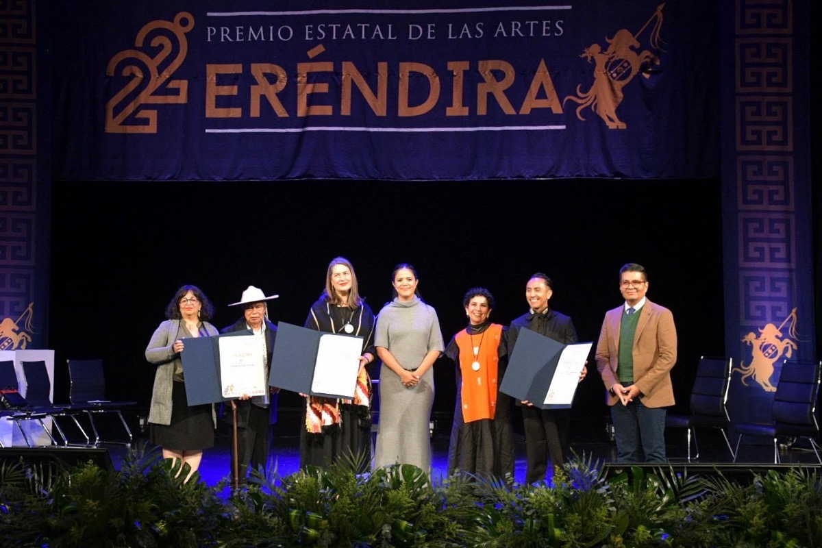 Un grupo de siete personas posa en un escenario con diplomas frente a un cartel que dice “Premio Estatal de las Artes Eréndira”.