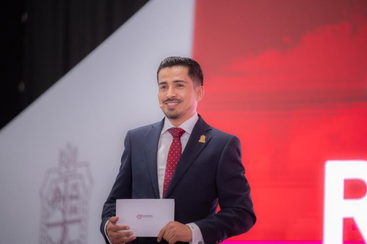 Un hombre con traje y corbata roja sostiene una tarjeta mientras sonríe en un escenario con fondo rojo y blanco.