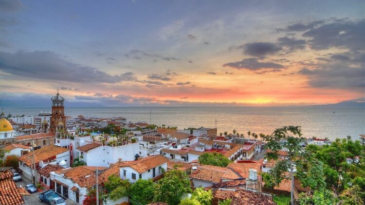 Vista de un pueblo costero con casas de techos rojos y el mar al fondo durante el atardecer.